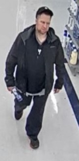 Caucasian man walking through a retail store, wearing a black baseball cap, dark sweater, dark jacket, jeans and black shoes.